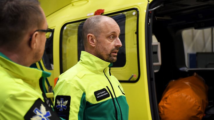 En man i en gr&ouml;n och gul uniform som st&aring;r bredvid en gul ambulans.