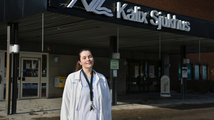 En kvinna i sjukv&aring;rdskl&auml;der som st&aring;r framf&ouml;r en byggnad.