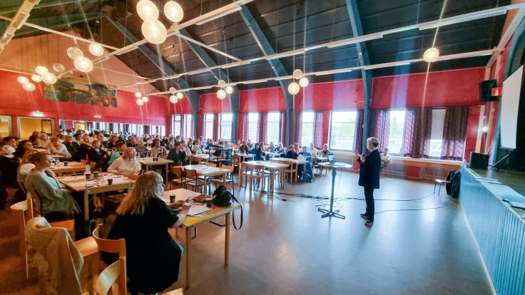 Åhörare i en samlingslokal, vända mot en föreläsare.
