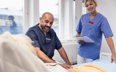 Sängliggande patient pratar med manlig sjuksköterska som sitter och kvinnlig läkare som står.