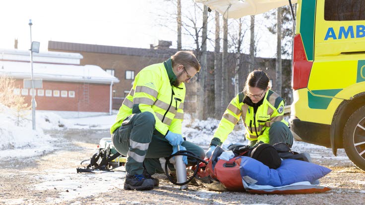 Två ambulanssjuksköterskor vårdar en patient, på en bår på marken, bredvid en ambulans.