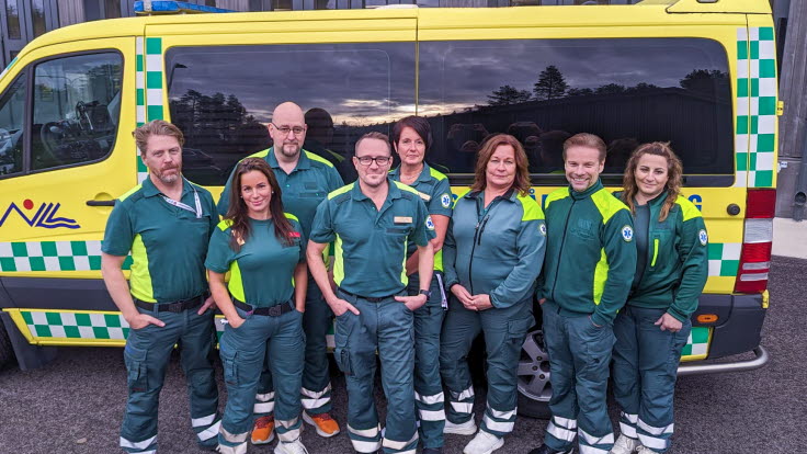 Åtta grönklädda sjuksköterskor framför en gul ambulans.
