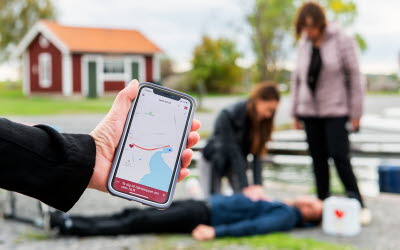 Mobil med appen Sms-livräddare och pågående hjärt- och lungräddning i bakgrunden.